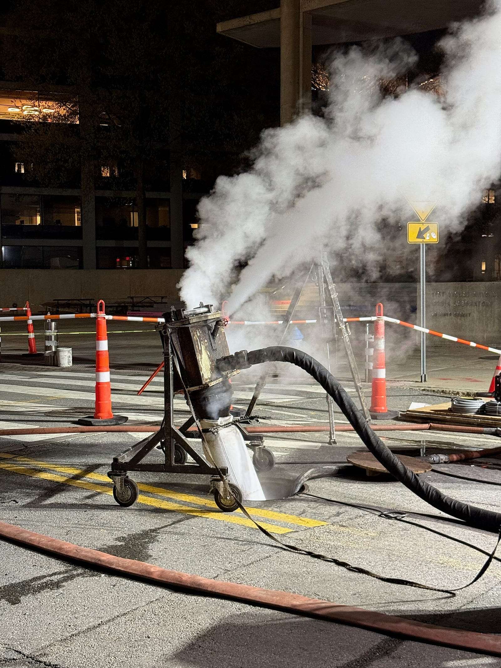 Cured-in-place pipe lining in progress, with steam and hoses running into a sewer access point on a city street